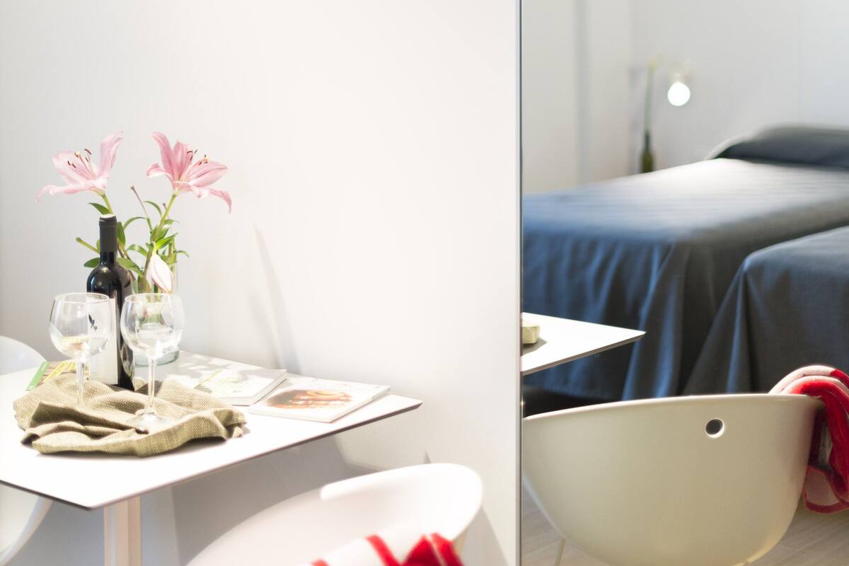 A modern hotel room with a bed, a small table with a vase of flowers, a chair, and a bedside table.
