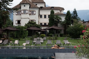 A pool area with lounge chairs and umbrellas, a large building with a tiled roof, and greenery in the background.
