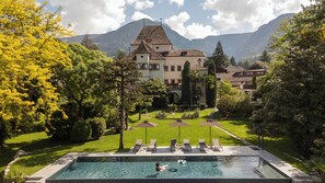 A pool with lounge chairs, a child in the water, and a large building with a mountain backdrop. - Castel Rundegg (Merano)