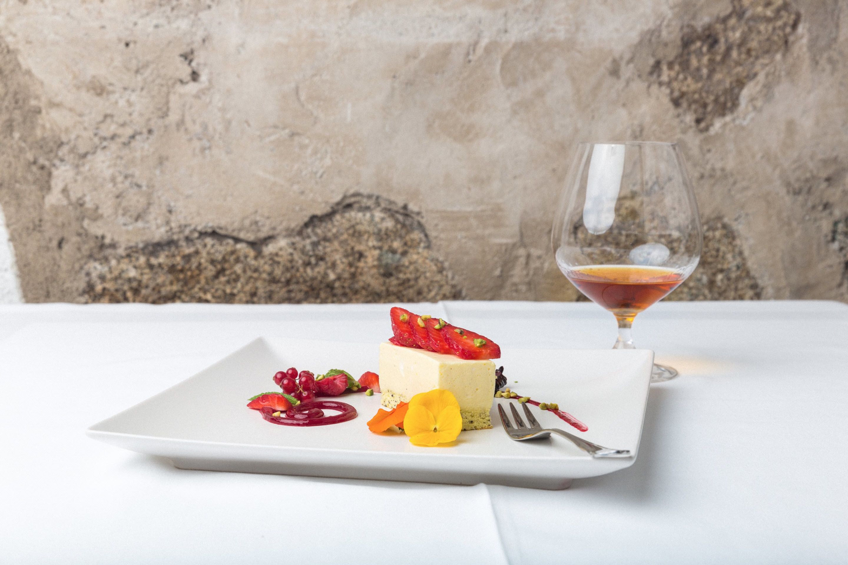 A dessert plate with a slice of cake, strawberries, and a glass of red wine on a white tablecloth.