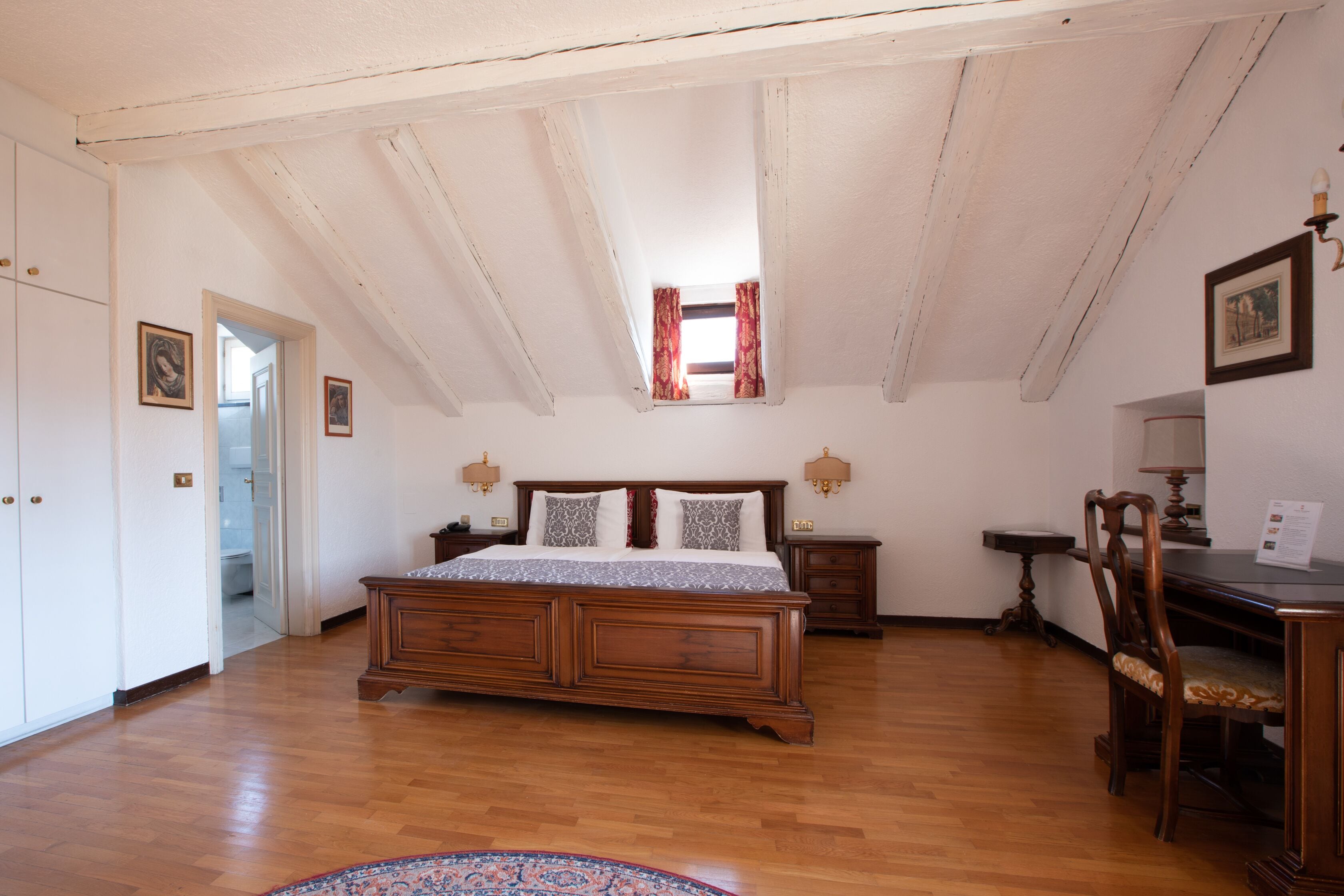 A bedroom with a wooden bed, a desk, a chair, a nightstand, and a small window with curtains.