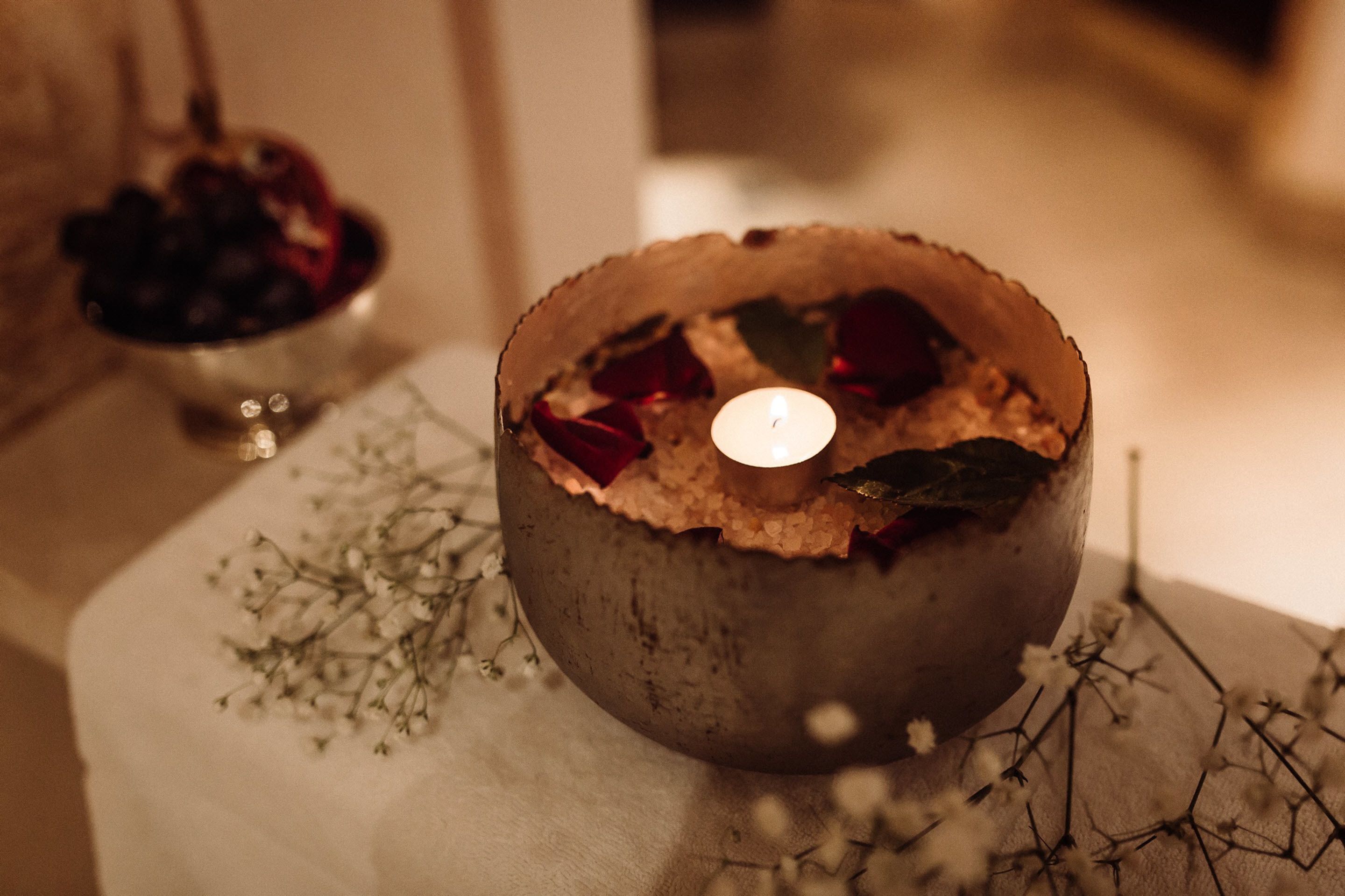 Eine Kerze in einer rustikalen Schale mit getrockneten Blumen und Beeren, platziert auf einem Tisch mit weißem Tuch und kleinen weißen Blüten.