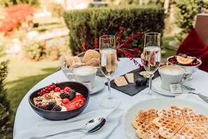 A table set outdoors with a bowl of mixed berries, waffles, and two glasses of a clear beverage, likely champagne, with a cup of coffee and a small plate of cheese.