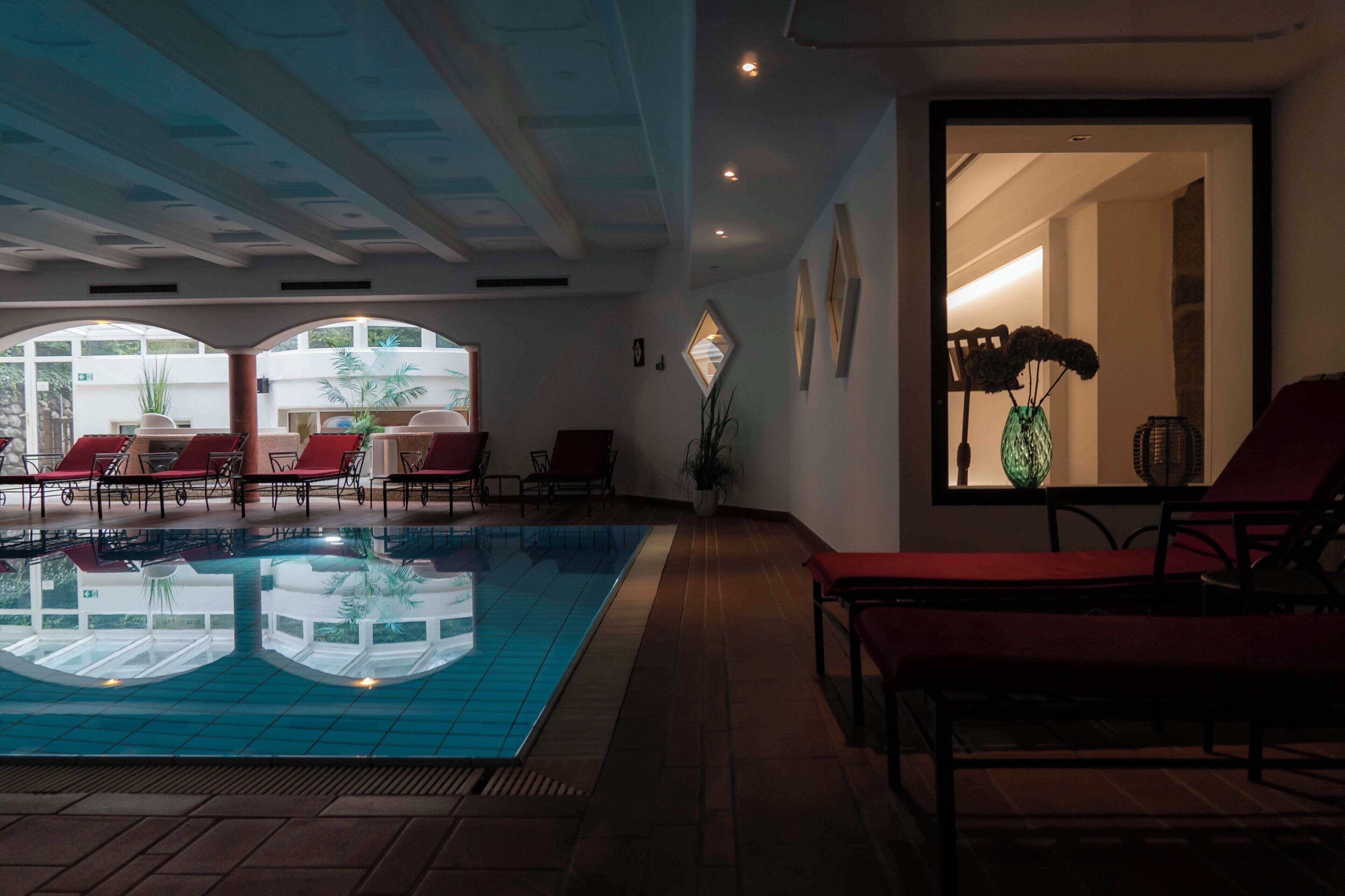 An indoor swimming pool with red lounge chairs, a potted plant, and a view of the outside through large windows.