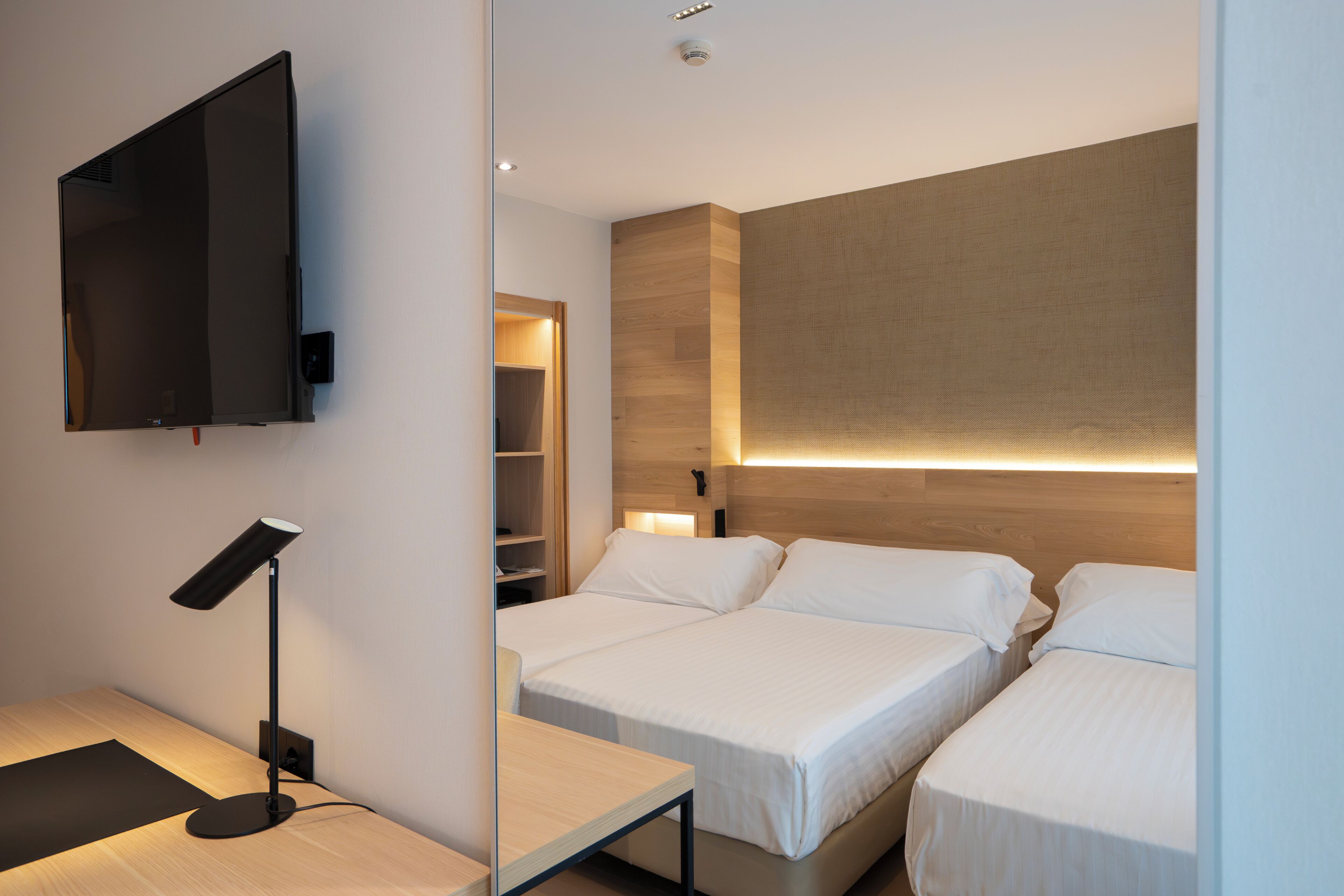 A modern hotel room with two beds, a desk, a TV, and a bedside table with a lamp.