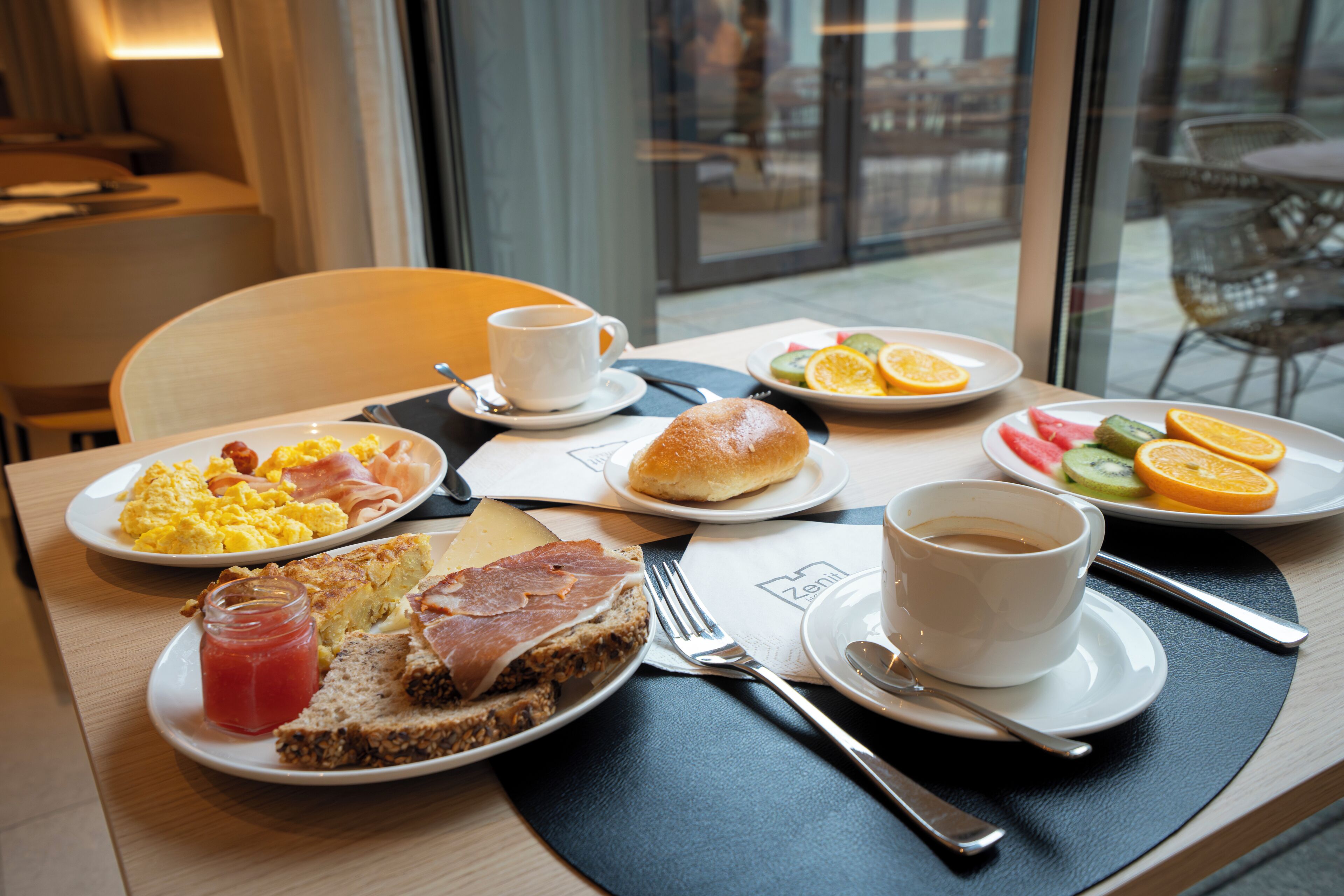 Una mesa de desayuno con platos de huevos, tocino, tostadas y fruta, junto con tazas de café y té.