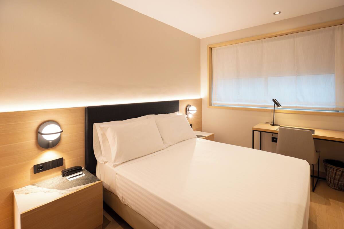 Une chambre d’hôtel bien rangée, avec un lit, une table de chevet, une lampe et un bureau avec une chaise.