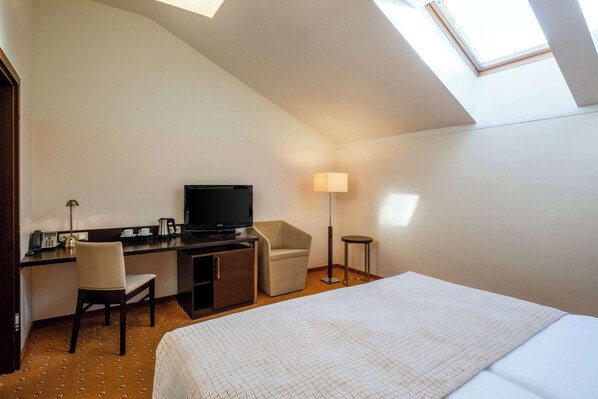 A hotel room with a bed, a desk with a television, a chair, a lamp, and a small table.