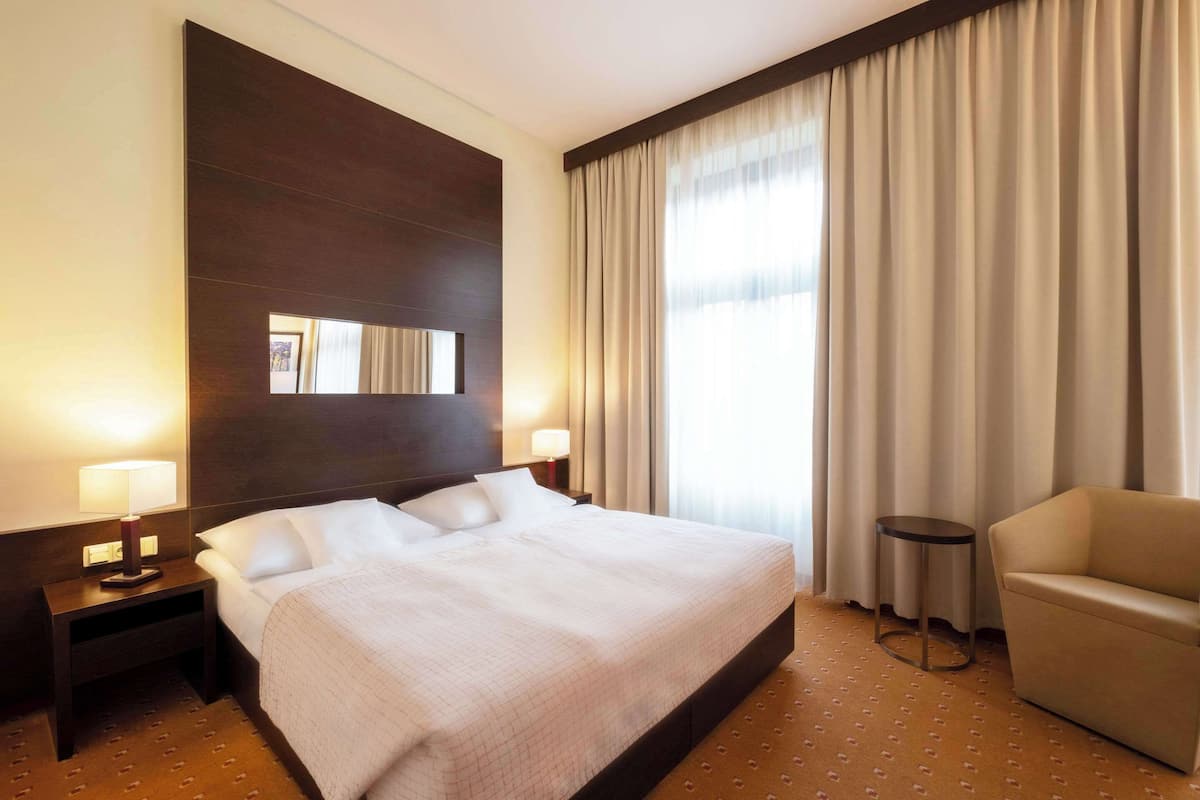 Ein Hotelzimmer mit einem großen Bett, einem kleinen runden Tisch, einer Couch und einem großen Fenster mit Vorhängen.