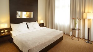 Una habitación de hotel con una cama grande, dos mesitas de noche con lámparas, un espejo en la pared y un florero con flores sobre una mesita auxiliar.