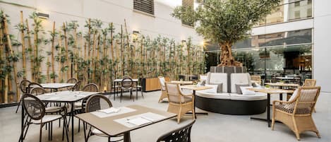 Un espace de restauration intérieur avec des murs en bambou, des tables rondes, des chaises en osier et un espace de repos central agrémenté d’un arbre.