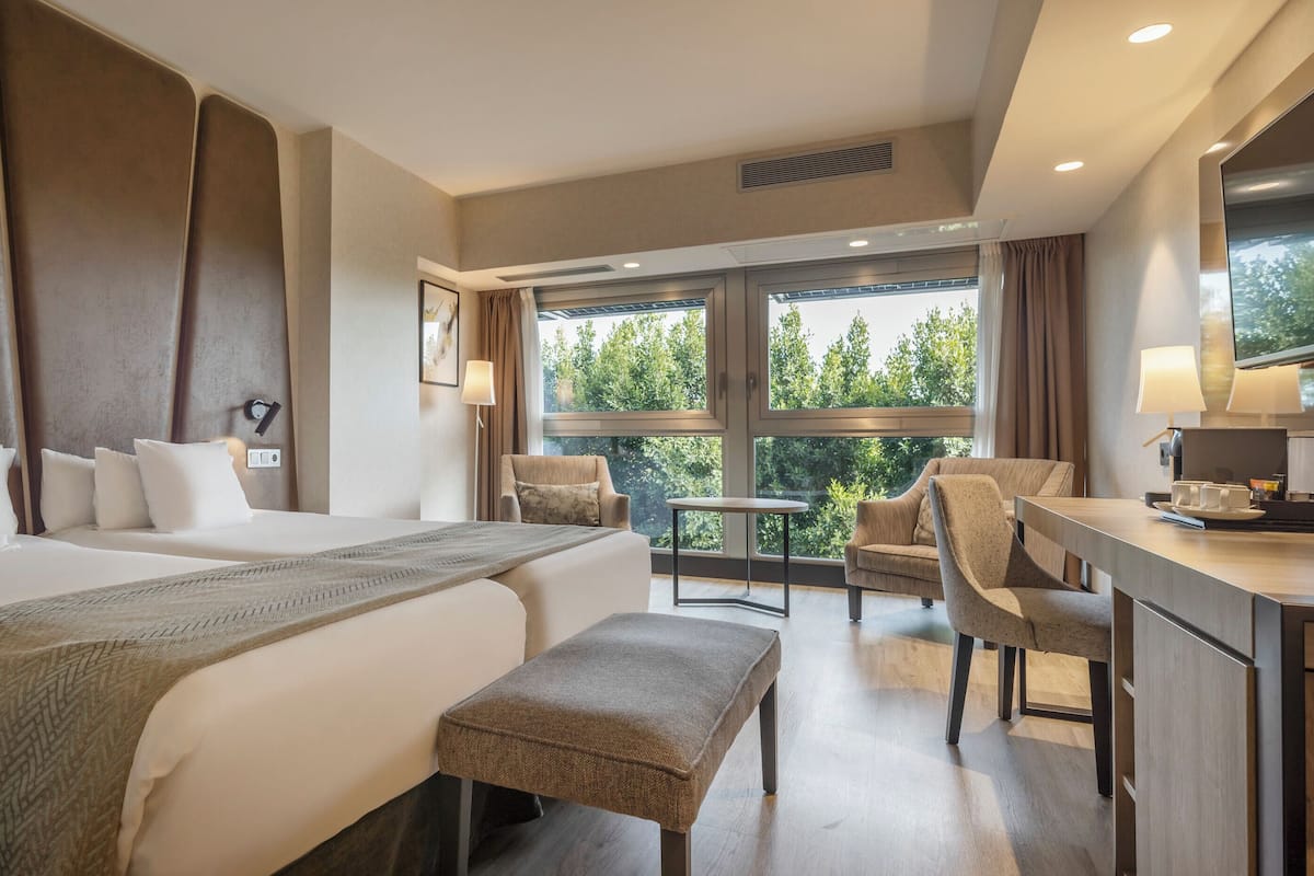 Habitación de hotel con dos camas, un escritorio, una silla y un pequeño balcón con vista a árboles.