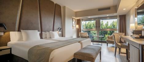 Habitación de hotel con dos camas, un escritorio, una silla y un pequeño balcón con vista a árboles.