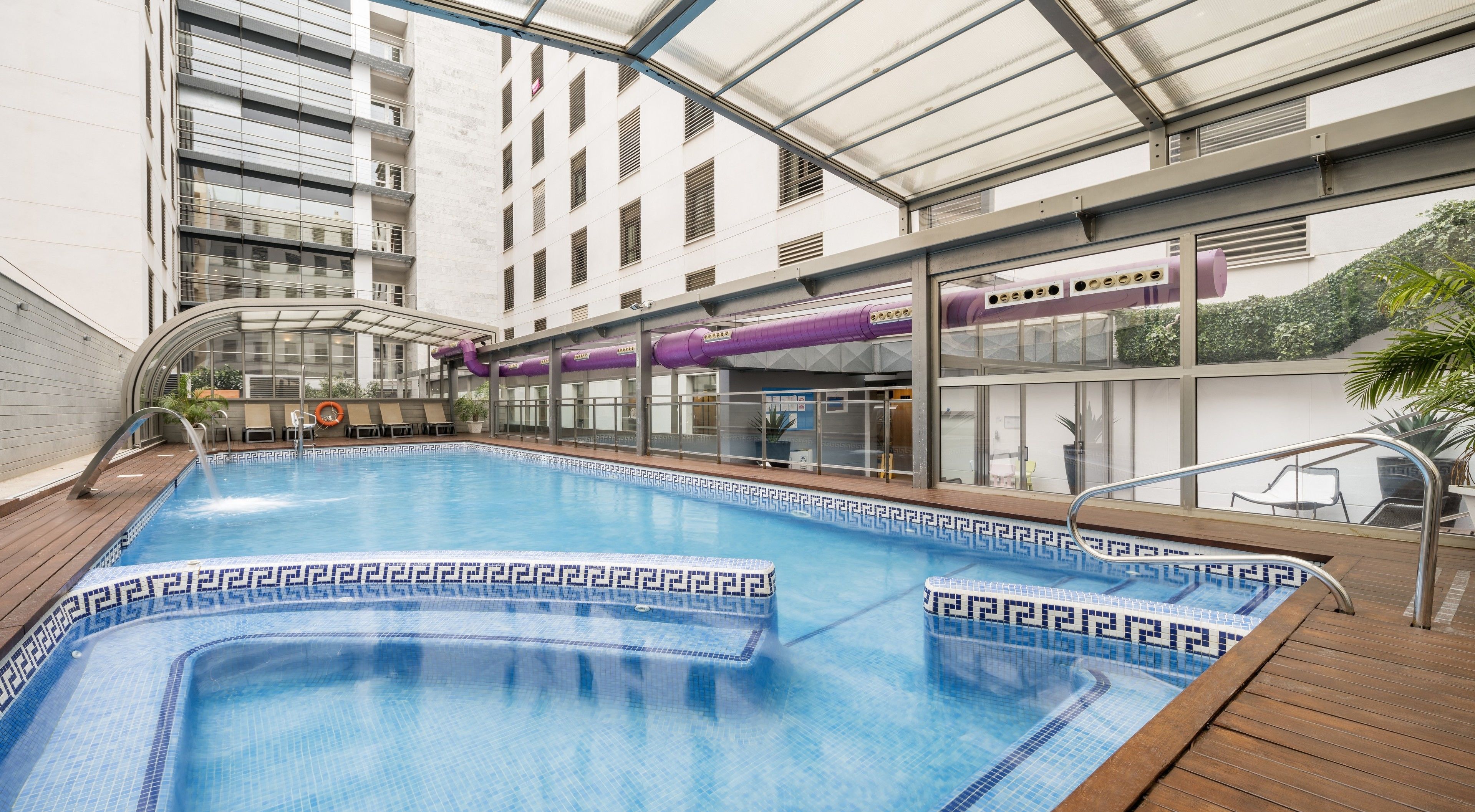 Una piscina interior con terraza de madera, paredes de vidrio y vistas del exterior del edificio.