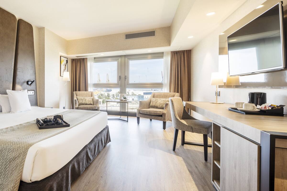 Habitación de hotel con una cama grande, un escritorio con silla, un comedor y un amplio ventanal con vistas.