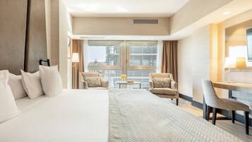 Een hotelkamer met een groot bed, twee fauteuils, een klein tafeltje met een lamp, een televisie en uitzicht op de stad door grote ramen.