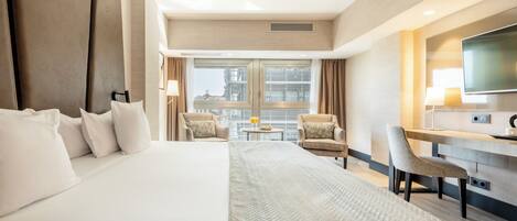 A hotel room with a large bed, two armchairs, a small table with a lamp, a TV, and a view of the city through large windows.