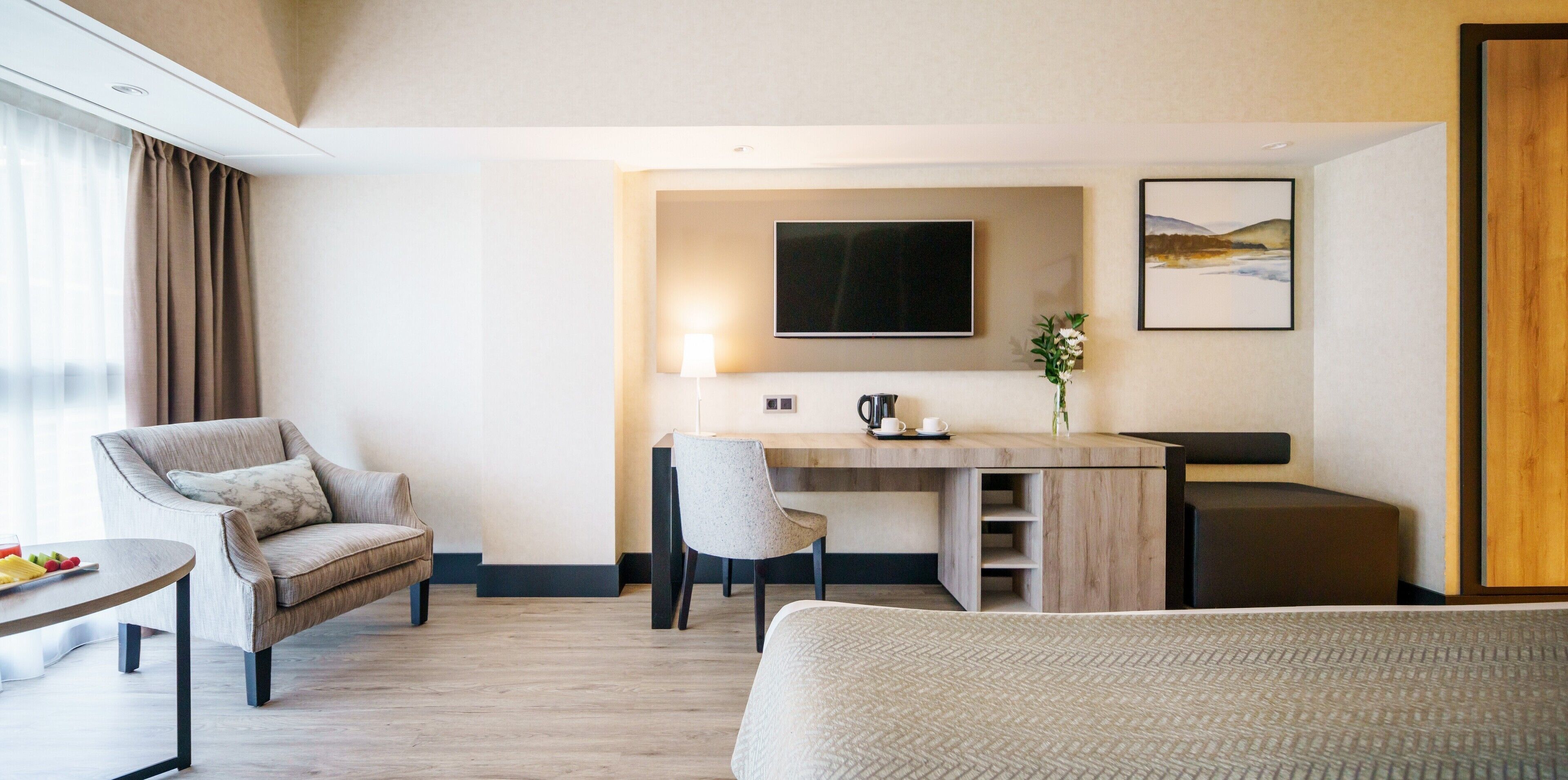 A modern hotel room with a bed, a desk, a chair, a TV, and a painting on the wall.