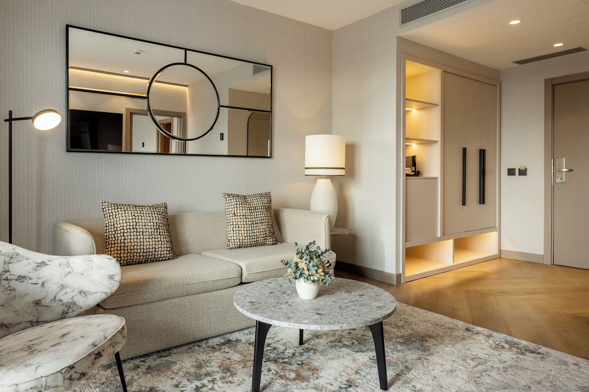 Una sala de estar moderna con sofá, sillón y una mesa redonda de centro. Un espejo grande y una lámpara de pie están fijados a la pared.