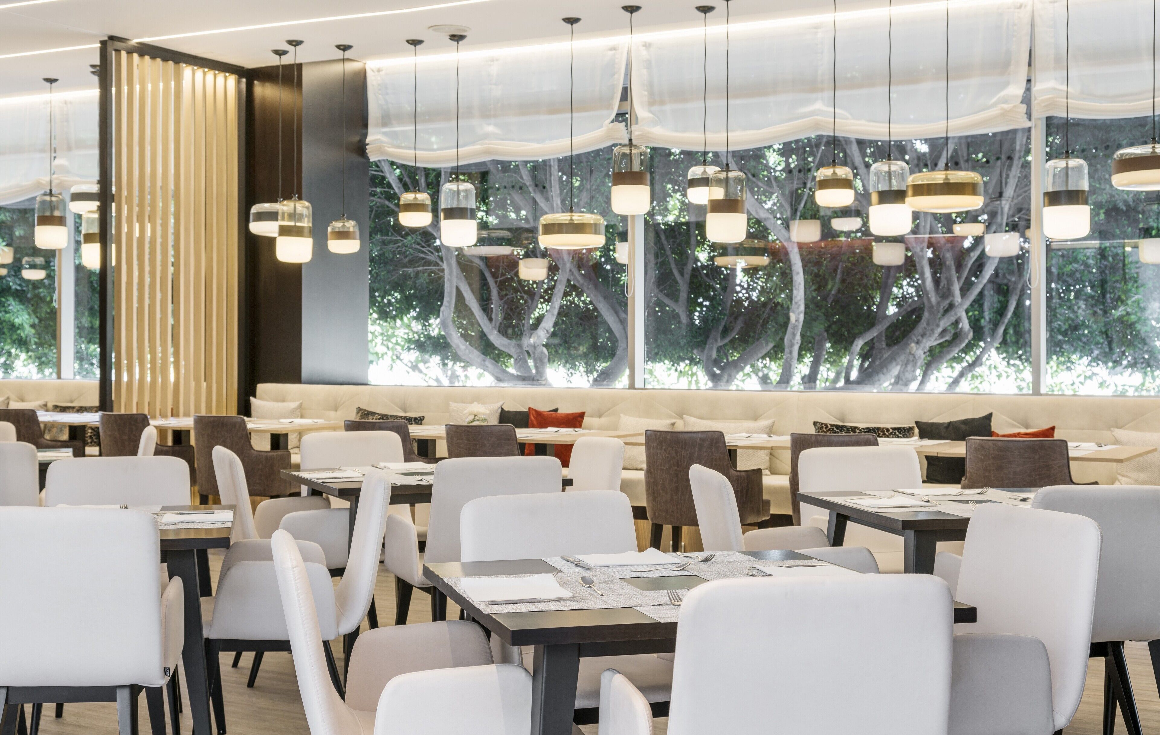 Um restaurante moderno com cadeiras e mesas brancas, luminárias pendentes e uma janela grande com vista para árvores.