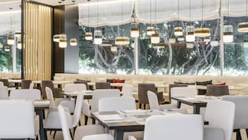 Een modern restaurant met witte stoelen en tafels, hangende pendantlampen en een groot raam met uitzicht op bomen.