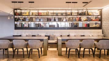 Ein modernes Wohnzimmer mit Sofa, Esstisch und Stühlen. Ein Bücherregal zeigt verschiedene Gegenstände, eine Deckenleuchte sorgt für Licht.