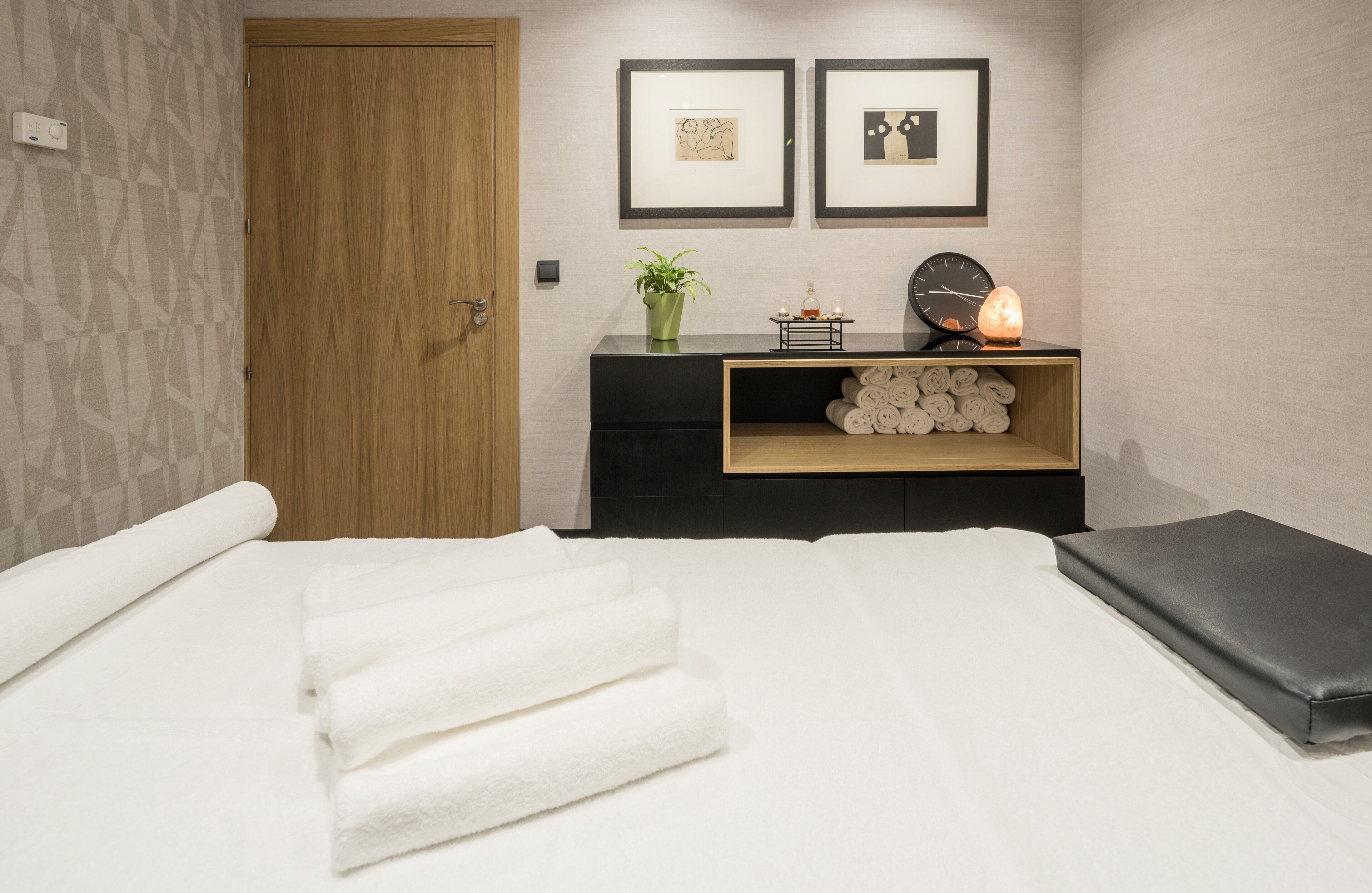 Habitación de hotel con cama, una puerta de madera, una mesita de noche negra con una planta, cuadros enmarcados y un reloj.