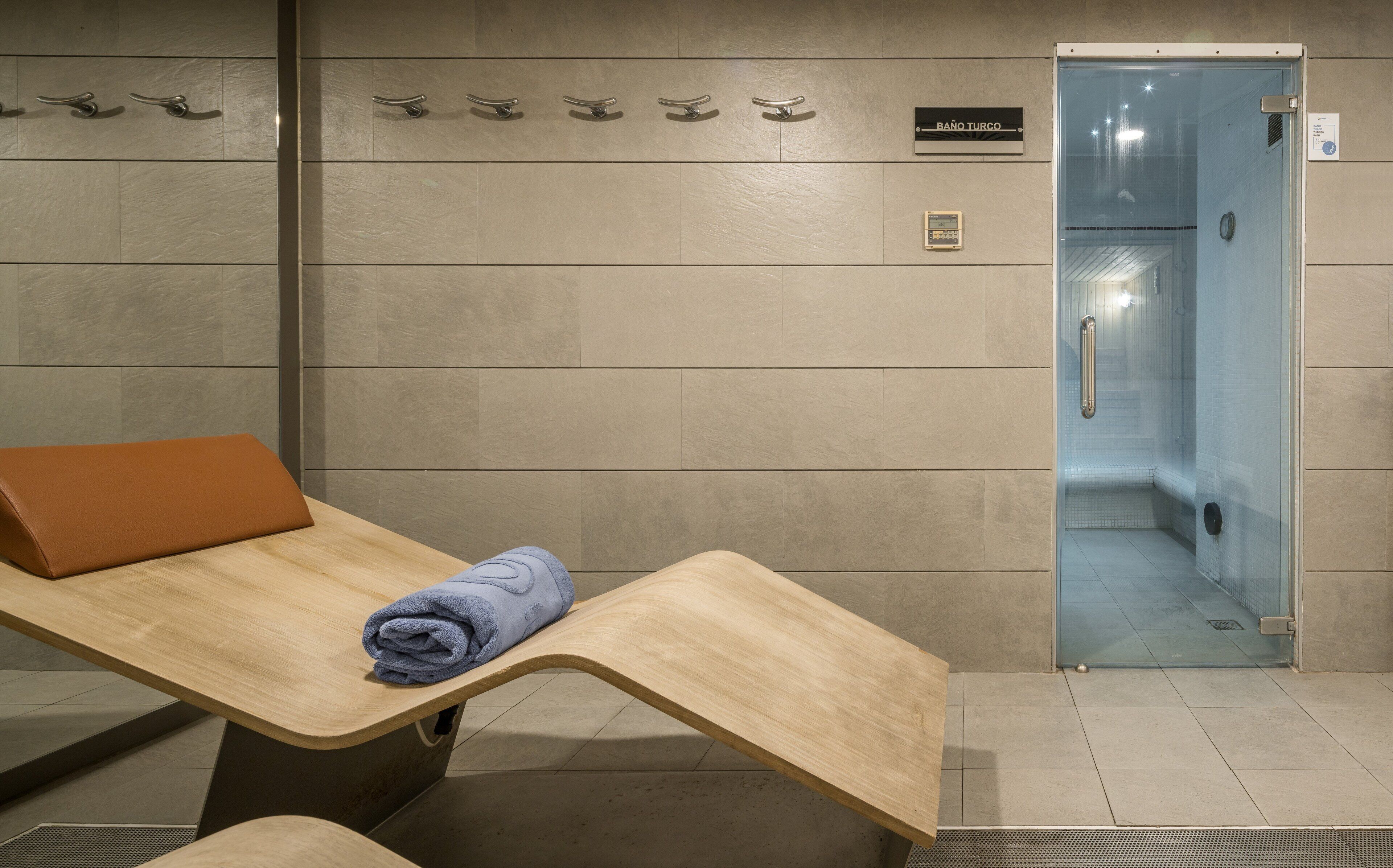 Un baño con un banco de madera, una toalla, una puerta de ducha de cristal y un letrero que dice "BAÑO TURCO".