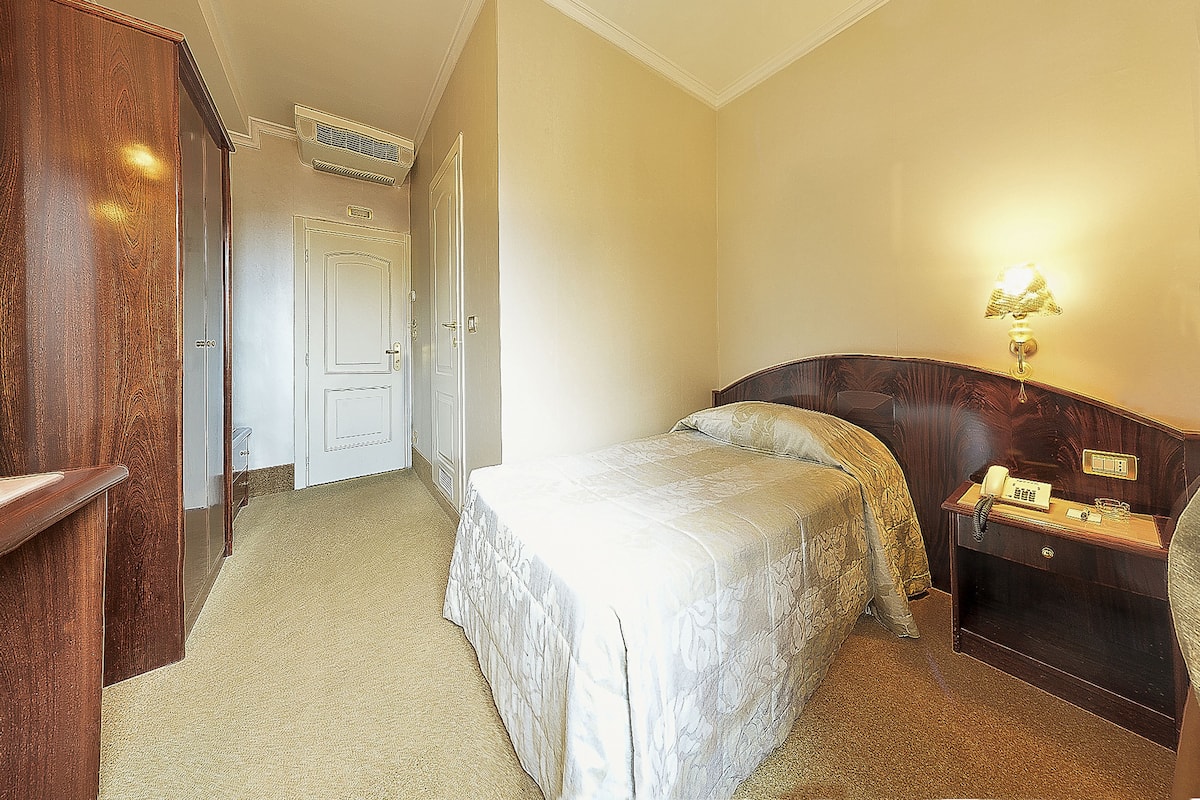 Una habitación de hotel con cama, mesita de noche con lámpara y un armario de madera.