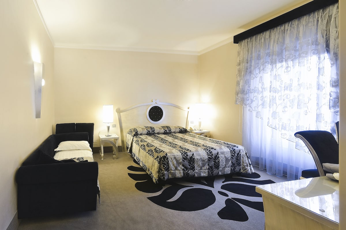 Habitación de hotel con cama, escritorio, silla, dos lámparas de noche y ventana con cortinas translúcidas.