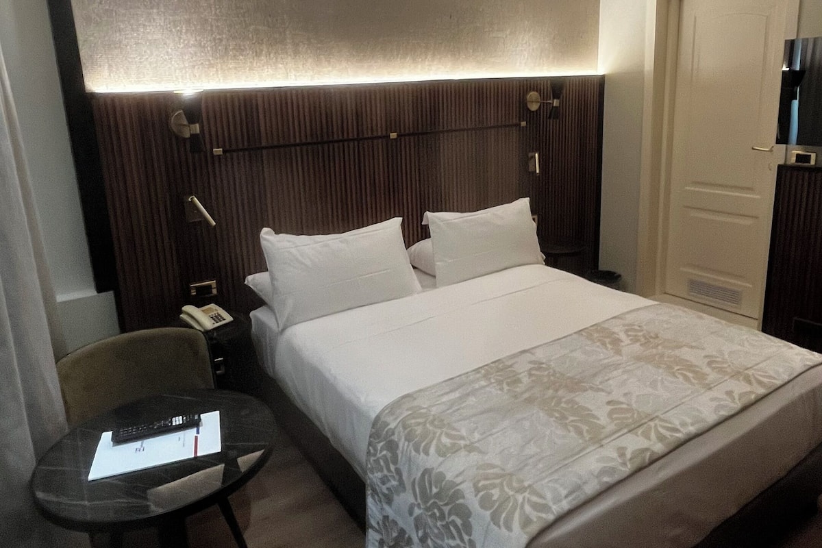 Habitación de hotel con una cama grande, una mesita de noche con lámpara, una silla y una pequeña mesa con control remoto.
