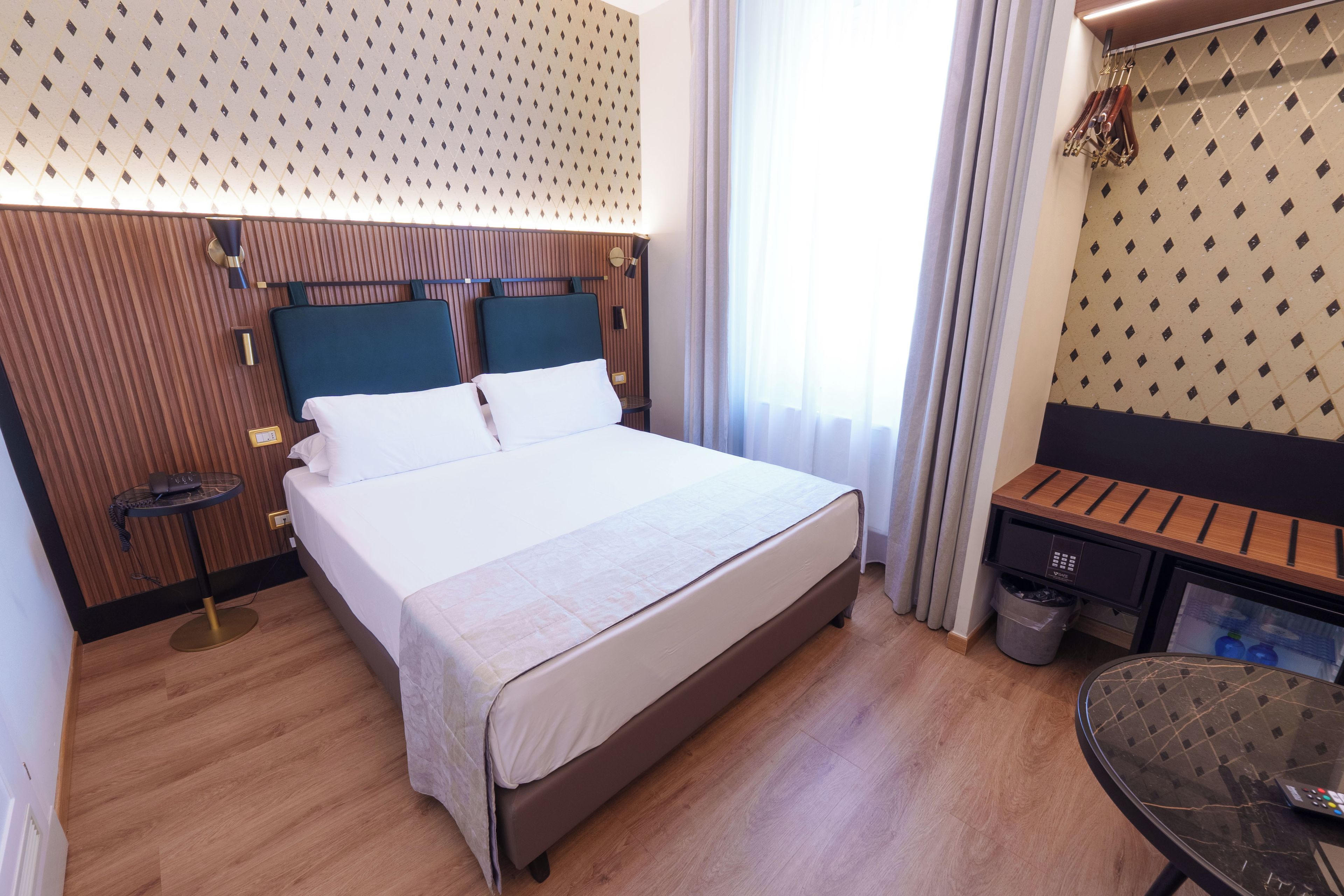 A hotel room with a large bed, a bedside table, a small desk, a chair, a trash bin, and a minibar.