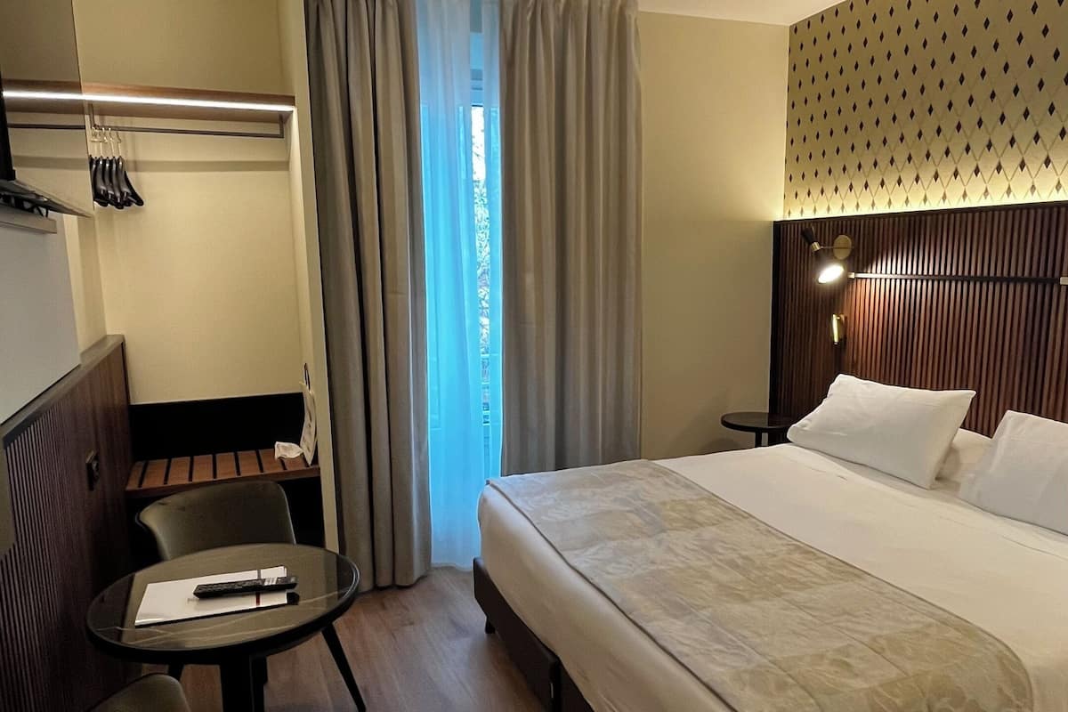 Una habitación de hotel con cama, escritorio, silla y ventana con cortinas.
