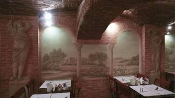 A dining area with brick walls, arched ceilings, and frescoes depicting classical figures. Tables are set with white tablecloths and cutlery.