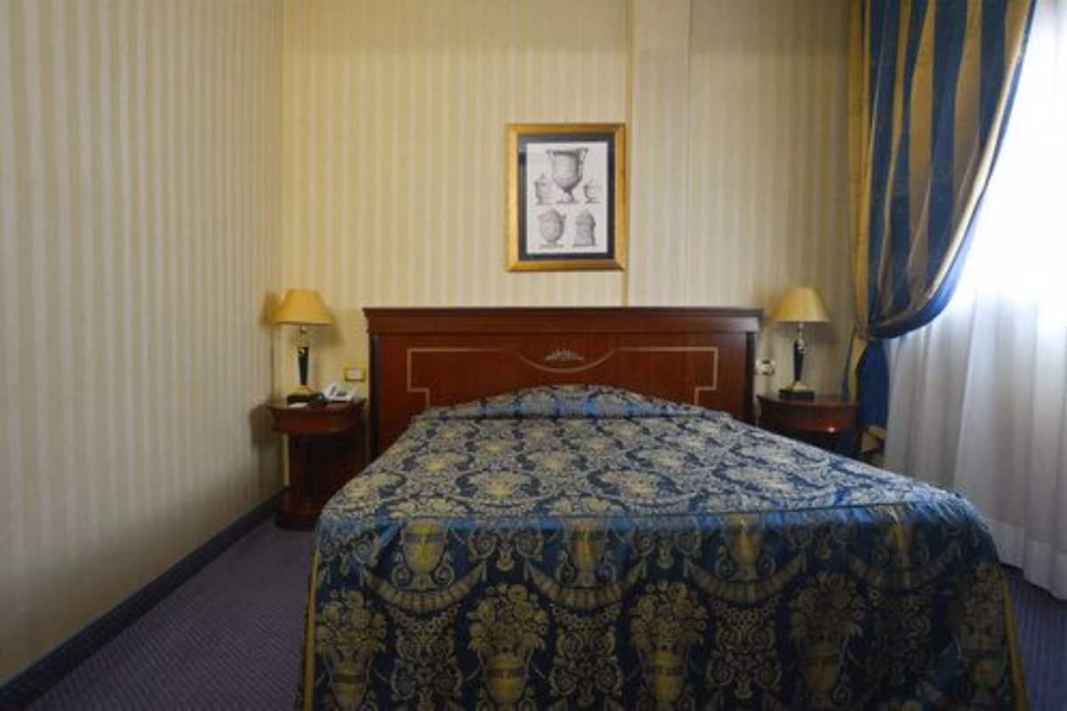 Habitación de hotel con una cama, dos mesitas de noche, un cuadro enmarcado en la pared y una ventana con cortinas.