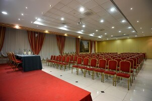 Uma sala de conferências com fileiras de cadeiras vermelhas, um palco com uma mesa e um projetor no teto.
