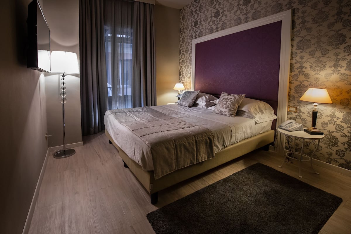 Habitación de hotel con cama, dos lámparas de noche, una mesita con teléfono y un ventanal con cortinas.