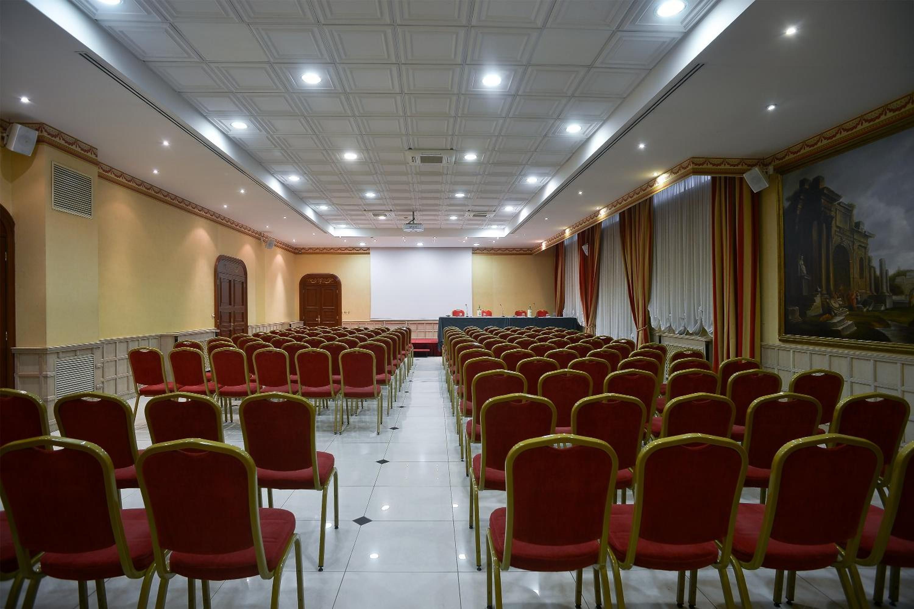 Ein Konferenzraum mit Reihen roter Stühle, einer Bühne und einem großen Gemälde an der Wand.