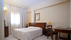 Ein Hotelzimmer mit einem Einzelbett, einem Schreibtisch, zwei Lampen, einem Sessel und einem gerahmten Bild an der Wand.