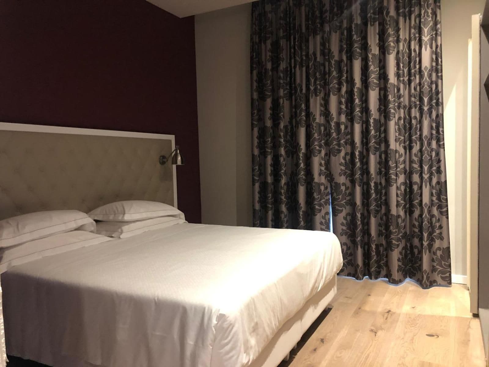 A hotel room with a bed, a headboard, a nightstand, and a window with curtains.