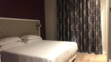 Una habitación de hotel con cama, cabecera, mesita de noche y ventana con cortinas.