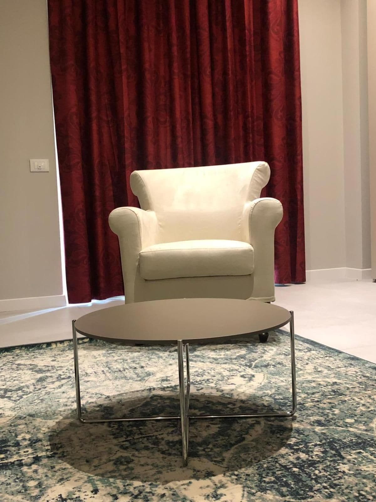 A white armchair with a round table in front of it, set against a red curtain backdrop.