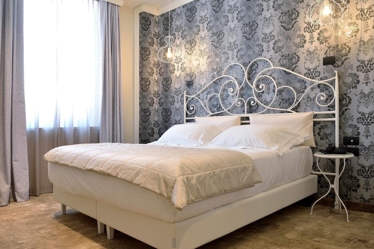 A neatly made bed with a white headboard and footboard, a bedside table with a lamp, and a patterned wallpaper.