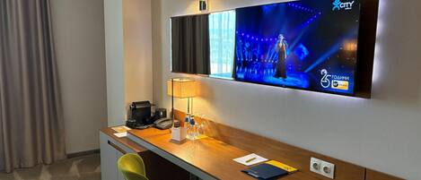 A hotel room with a desk, chair, TV, lamp, and a coffee maker.