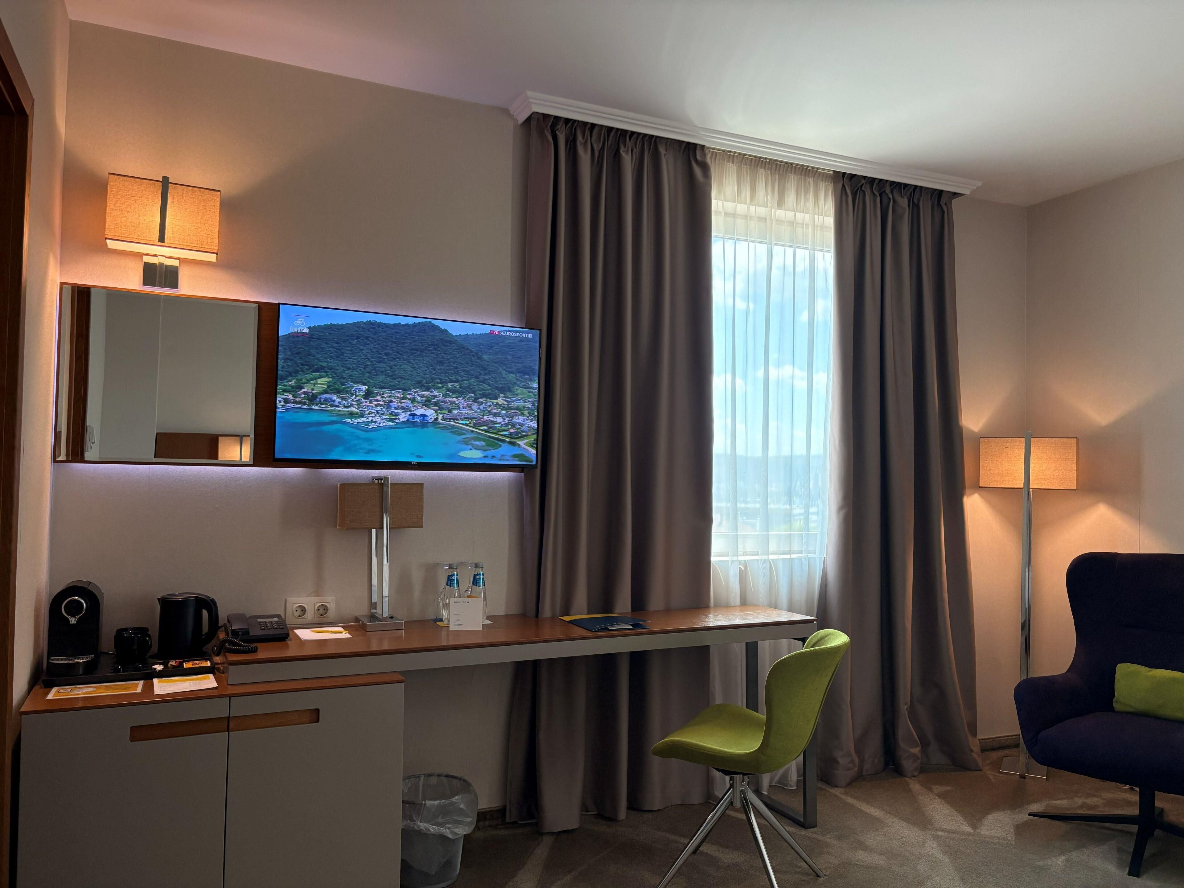 A hotel room with a desk, chair, TV, and a large window with curtains.