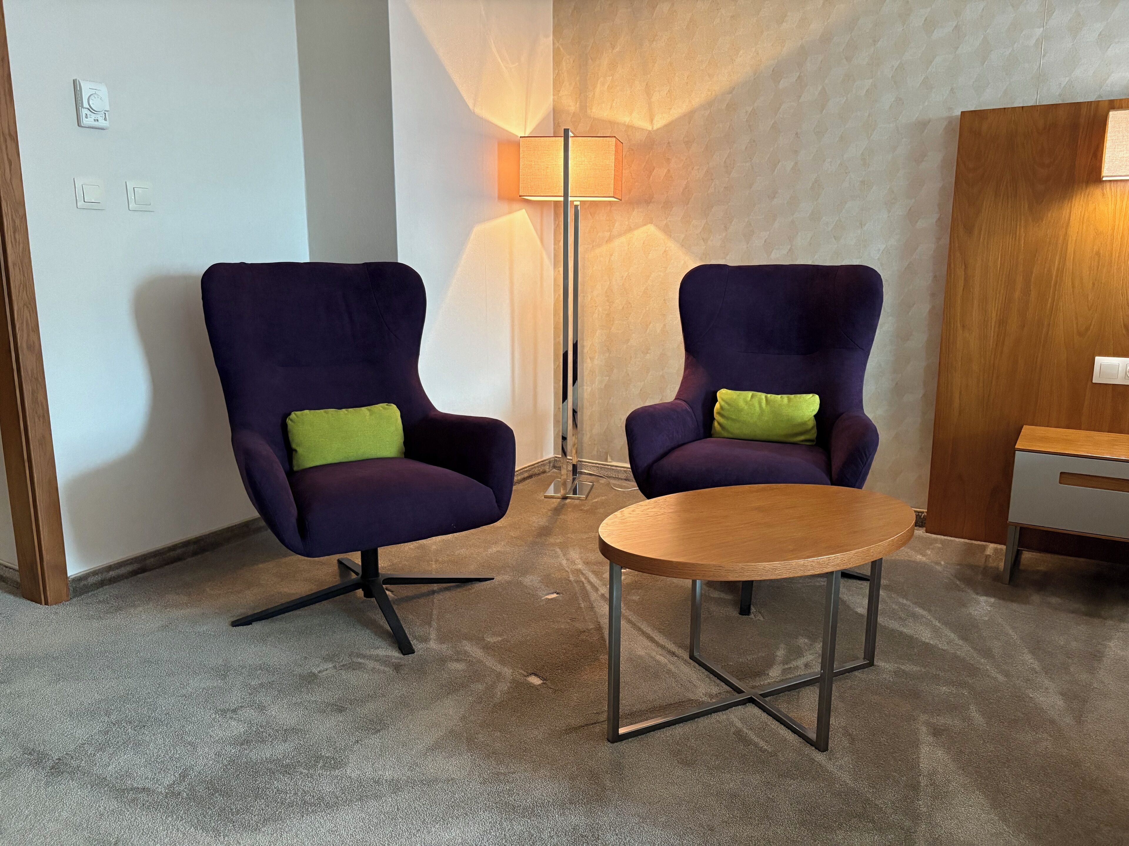 Dos sillones morados con cojines verdes, una mesita de madera, una lámpara de pie y una puerta de madera.