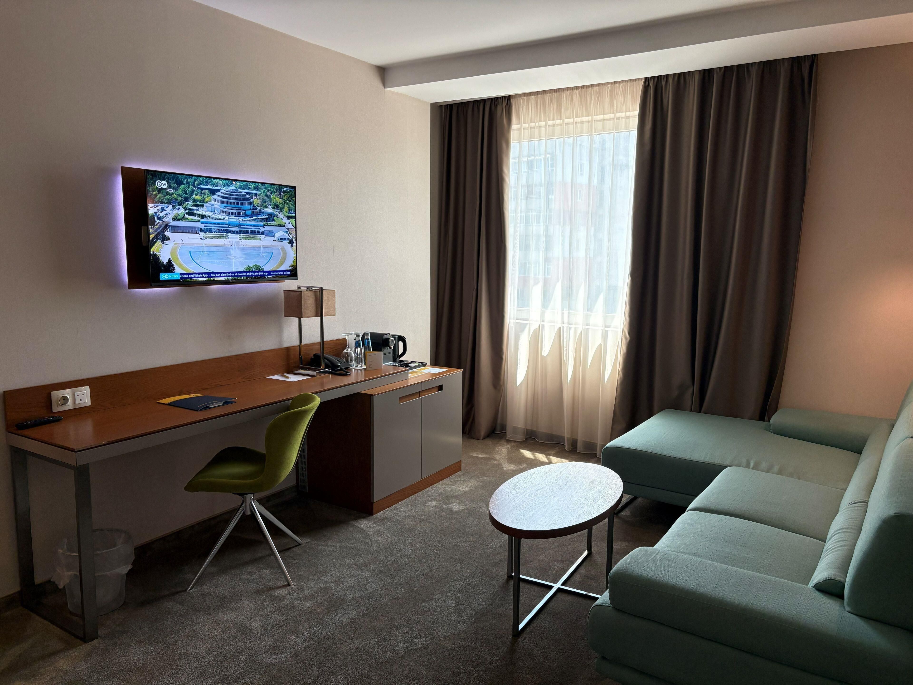 A hotel room with a desk, a green chair, a small round table, a sofa, a TV mounted on the wall, and a window with curtains.