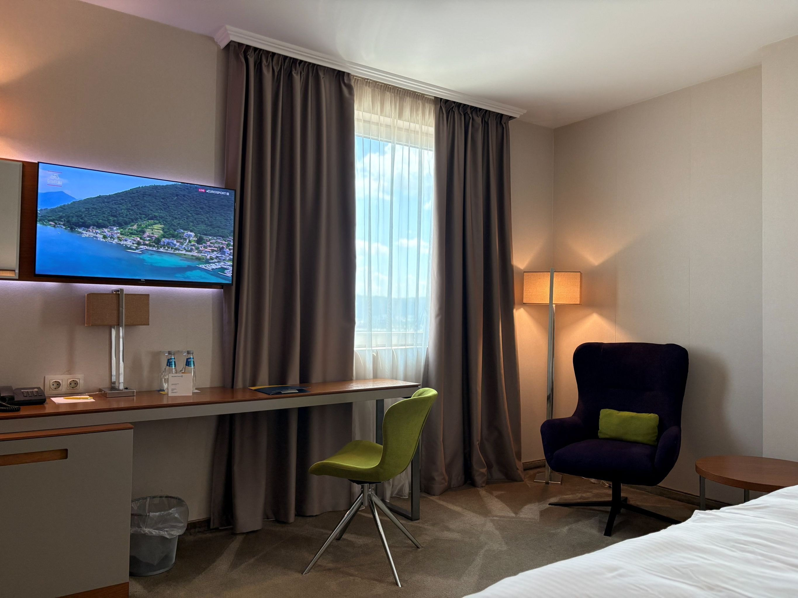 Une chambre d’hôtel comprenant un lit, un bureau avec un ordinateur, une chaise, un téléviseur diffusant des images de paysages et une lampe.