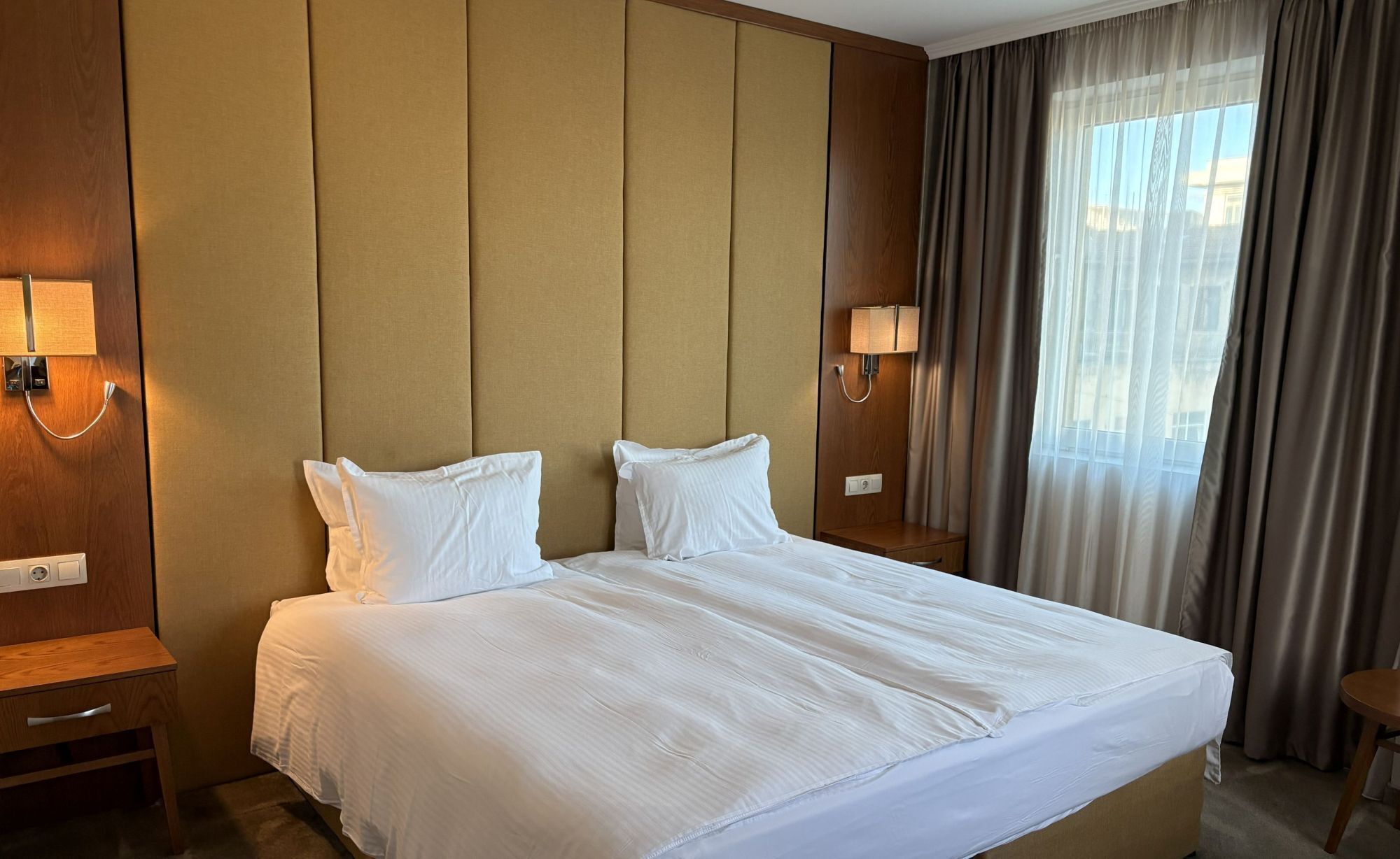 A hotel room with a large bed, two bedside tables, a nightstand, a lamp, and a window with curtains.