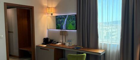 A hotel room with a desk, chair, TV, and a window with curtains.